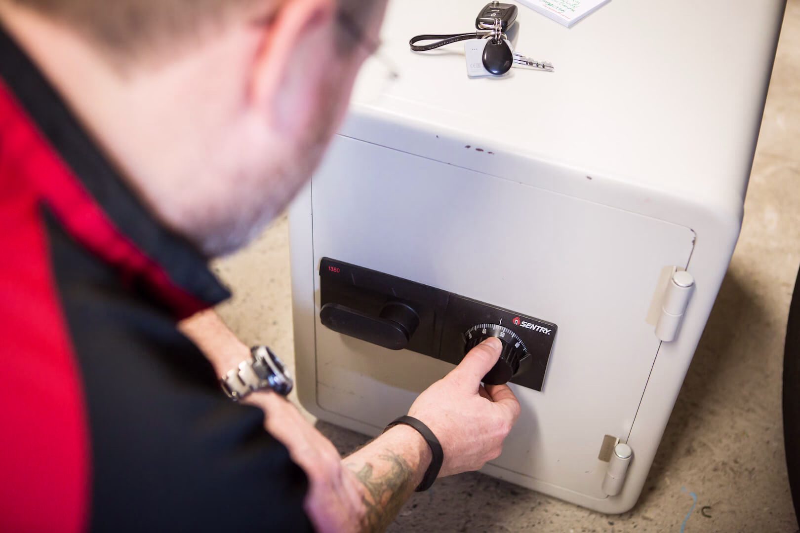 Expert Locksmith Unlocking a Secure Safe A locksmith's hands working on a metallic safe, turning a combination lock to unlock it. The safe door is partially open, revealing a dark interior. Tools like a stethoscope and lockpicking instruments are visible nearby, set against a plain workbench background.